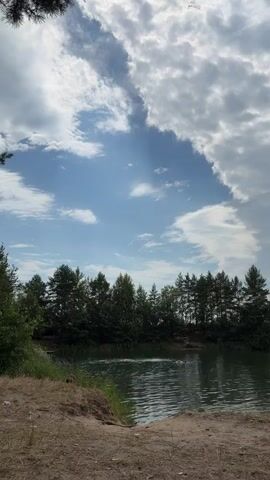 The video shows a natural scene with a lake surrounded by trees. The sky is partly cloudy, with sunbeams breaking through the clouds. A few people are visible in the water, appearing to be swimming or playing. The foreground features dry grass and sand, suggesting a warm, possibly summer day.