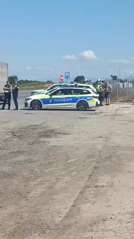 The video shows a traffic stop involving several police officers and two vehicles, a white van and a semi-trailer truck. Two police cars are parked behind the van and the truck. Three uniformed police officers are standing near the van, two of them are facing each other while one is looking in the direction of the truck. Another two uniformed police officers are standing near the trailer of the truck, facing the van. Another officer is standing to the side near the police car. The scene takes place on a paved road in a rural area with a clear blue sky and some trees and hills in the background. The van's rear doors are open, and it appears to be a daytime operation.