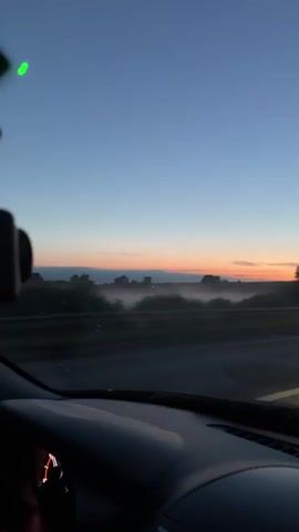 The video shows the interior of a car driving on a road at dusk. The dashboard and steering wheel are visible, along with a smartphone displaying the time "22:22". Outside the car, the sky is a gradient of deep blue and fading orange, suggesting sunrise or sunset. A thin layer of fog or mist hovers over a treeline in the distance. The overall atmosphere is calm and serene, with the soft light and quiet hum of the car creating a sense of peaceful travel. The location is identified as Malaya Ven'ya, Russia.