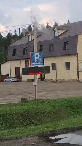 The video shows a parking lot in front of a building. A sign with a blue "P" and the word "ОХРАНЯЕМАЯ" (meaning "guarded" or "protected" in Russian) is mounted on a pole. A silver car is parked to the left of the building. A person is sitting on a bench in front of the building. The environment appears to be rural with trees in the background. There is some greenery and a puddle in the foreground. The sky is cloudy.