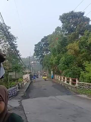 The video shows a road with several motorbikes. The road appears to be paved and has guardrails on both sides. There are trees and vegetation on the right side of the road, and buildings on the left. The sky is overcast. In the foreground, a person wearing a hijab and a dark jacket is visible on the left side of the frame. In the background, several people are riding motorbikes. One motorbike in the center of the frame has its headlight on. Another motorbike is seen further up the road. The video appears to be taken from a moving vehicle.