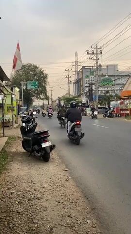 A cloudy sky looms over a street with a mix of residential and commercial buildings. A road runs through the center of the frame, with several motorcycles driving on it. On the left side of the road, there is a row of shops and a dirt path lined with parked motorcycles. A flag, likely the Indonesian flag due to its red and white colors, is flying on a pole in front of one of the buildings. A tall electrical tower is visible in the background, along with power lines crisscrossing the sky. The overall scene appears to be a typical street in a populated area, possibly a town or city.