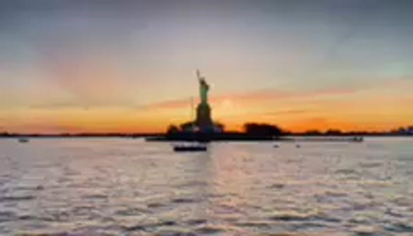 This media file captures a serene, wide shot of the Statue of Liberty at sunset, viewed from the water. The iconic monument stands silhouetted against a vibrant sky painted with hues of orange, yellow, and purple, indicative of a setting sun. Rays of light emanate from behind the statue, adding a dramatic flair to the scene. The water in the foreground is calm, reflecting the colors of the sky and creating a shimmering surface. Several small boats are scattered across the bay, some in the middle distance and others closer to the viewer, adding a sense of scale and activity to the otherwise tranquil setting. The distant shoreline is also visible as a dark silhouette. The overall mood is peaceful and majestic, highlighting the grandeur of the monument and the beauty of the natural light. There is no visible text in the media file.