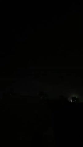 The video begins with a black screen, then shows a dark night sky with faint clouds. The camera pans to reveal a palm tree with dried fronds and a small green light in the distance. A white car is partially visible on the right. The camera then moves to show a white porch with a railing, and more palm fronds. The scene is dimly lit and appears to be outdoors at night.