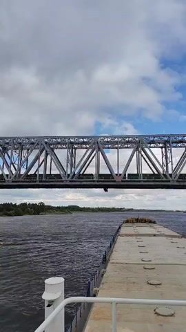The video shows a view from a boat looking towards a large metal train bridge crossing a wide river. The boat is moving slowly. The bridge is made of a truss structure and there is a train visible on top of it. The sky is cloudy and overcast. The riverbanks on either side are lined with trees. The boat appears to be a barge or cargo vessel, with part of its deck visible in the foreground.