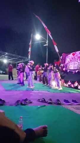 At night, a group of young people are dancing on a green carpet in front of a stage. They are wearing casual clothes, including t-shirts, jeans, and hats. Some of them are wearing headscarves. There are Indonesian flags on poles behind them, and a banner with text in Indonesian on the stage. In the foreground, there are several pairs of shoes and a water bottle on the ground. The lighting is dim, with a spotlight illuminating the dancers. There are also some people watching the performance in the background.