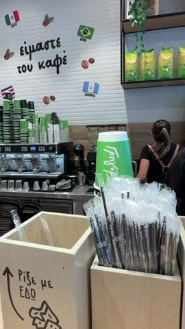 The video shows the interior of a coffee shop. In the foreground, there are wooden containers filled with plastic straws. A man in a black t-shirt and a baseball cap is standing behind the counter, looking towards the right. He is wearing brown leather suspenders. To the left of him, another person is partially visible behind a counter, also wearing a black t-shirt and suspenders, with their back to the camera. In the background, shelves are stocked with bags of coffee beans. There are also coffee machines and other equipment on the counter. The walls are white brick. There is some Greek writing on the wall, which translates to "we are the coffee" and "throw it here". The overall impression is a busy, operational coffee shop environment.