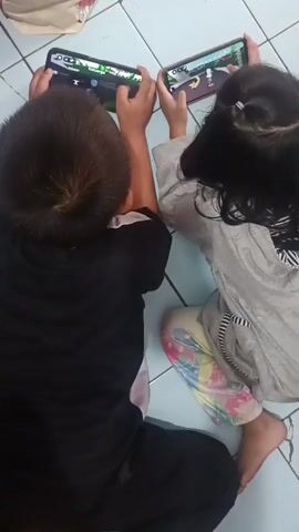 This media file shows two children, a boy and a girl, playing video games on their phones. They are both lying on their stomachs on a tiled floor, with their phones held horizontally in front of them. The boy, on the left, is wearing a black t-shirt with a pink stripe. The girl, on the right, is wearing a gray hoodie and colorful patterned leggings. Both children appear engrossed in their games, their faces tilted towards the screens.

The scene is an indoor setting, possibly a home, as indicated by the tiled floor and the presence of what appears to be a rug or blanket in the top left corner. The lighting suggests it is daytime, though no specific time can be determined. The children's focus and the nature of the activity suggest a casual and leisurely moment. There is no discernible text visible in the media file. The overall atmosphere is one of focused entertainment.