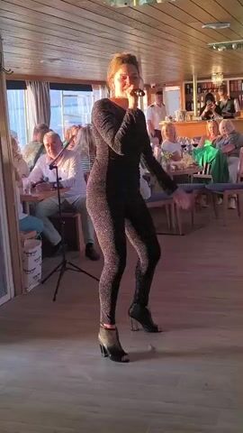 A woman wearing a sparkling black jumpsuit and open-toed heels sings into a microphone while standing on a wooden floor. She is positioned in the foreground of the video, facing forward with her body slightly angled to the left. Behind her, in a seated area, several people are visible at tables. The room appears to be a dining or lounge area with windows showing an outdoor scene. The lighting suggests it might be late afternoon or early evening.