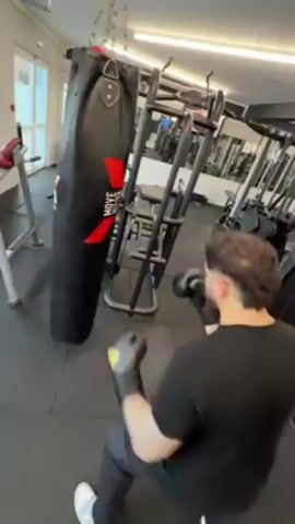 A man in black sportswear is hitting a black heavy bag with red accents. The bag is suspended by chains in a fitness center, which includes various weightlifting machines and other equipment. The man is wearing boxing gloves and throws punches at the bag repeatedly. The video is shot in a first-person perspective, handheld style.