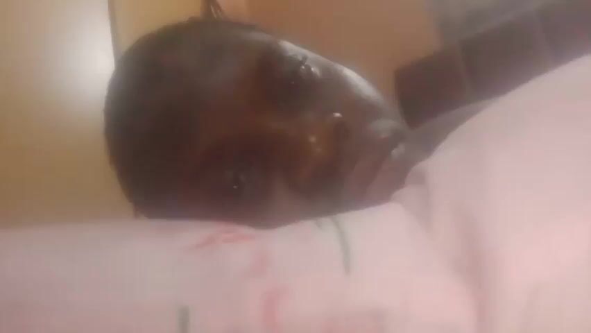 A person with dark skin is lying on a pink patterned pillow. The person is looking to the right. A brown cabinet is in the background. The lighting is dim.