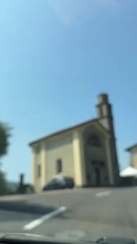 A car drives past a stone church with a bell tower on a sunny day. The church has a light-colored facade and a dark-tiled roof. A car is parked in front of the church. Further down the road, a building with the words "CASA ROSA" painted on its side is visible. The surrounding environment appears to be a small town or village.