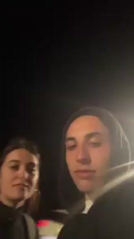 Two young adults are visible in the video frame. A woman with dark hair pulled back is on the left, and a person in a hooded sweatshirt is on the right. The video is shot at night, and it appears they are outdoors, possibly near a street due to the presence of bright lights. Both are looking at the camera, talking and moving their mouths. The person in the hoodie appears to be making blowing motions. The camera view is close-up.