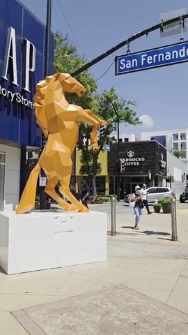 A geometric orange horse sculpture stands on a white pedestal in front of a GAP store and a Starbucks. A street sign reading "San Fernando" is visible above. People are walking on the sidewalk and cars are driving by in the background. The sky is blue and the sun is shining.