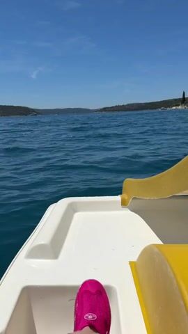 The video is shot from the perspective of someone on a pedal boat. The front of the pedal boat, including a yellow slide, is visible in the foreground. The person's feet wearing bright pink shoes are also visible at the bottom of the frame. The boat is on a body of water, likely the sea, with small waves. In the background, there is a coastline with green hills covered in trees. Several houses and buildings, some with red roofs, are situated along the coast. The sky is bright blue with a few wispy clouds. The overall scene depicts a sunny day at a coastal location.