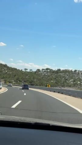 The video shows a first-person perspective from inside a car driving on a highway. The road curves to the right, with a mountainous, vegetated landscape on the left. Several cars are visible on the highway, including a blue car directly ahead. Yellow curve warning signs are placed along the right side of the road. In the distance, a tunnel entrance is visible on the left side of the highway, with a truck and other cars entering it. The sky is blue with scattered white clouds. The video appears to have been filmed during the day.