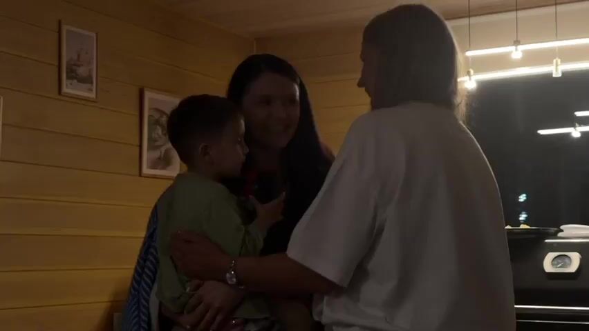 The video shows three people in a room with wood-paneled walls. In the center, a woman is holding a young boy. She is laughing and appears to be interacting with another woman standing to her right, who is wearing a white t-shirt. The boy is wearing a light green shirt and is looking down. On the left wall, there are two framed pictures. The room is lit by overhead lights. The overall mood seems joyful and familial. The timestamp suggests the video is very short, possibly a snippet of a longer recording. The context of Kubovaya, Russia, indicates a likely domestic setting.
