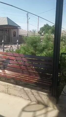 This is a short video of an outdoor scene. The camera pans to the right, showing a wooden bench in the foreground. Behind the bench is a decorative metal fence and some greenery, including small pine trees. In the background, there is a house with a gray roof and a gated entrance. The sky is clear and blue.