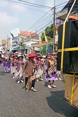 A street parade is underway, featuring a group of people in traditional attire. The participants, a mix of adults and possibly older children, are walking in formation down a paved street. They are mostly wearing conical hats, long-sleeved tops, and patterned skirts or sarongs. Some are also adorned with sashes and accessories. The atmosphere appears festive, with flags visible in the background and a large speaker system on wheels, suggesting music is accompanying the procession. The buildings lining the street suggest an urban or town setting. The sun is shining, indicating it is daytime.