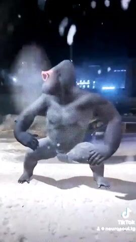 A CGI gorilla wearing pink heart-shaped sunglasses is dancing in a sandy area at night. The gorilla is in a squatting position and then stands up and moves its arms. There appear to be particles of snow or confetti falling around the gorilla. In the background, there are lights from buildings and a dark colored car.