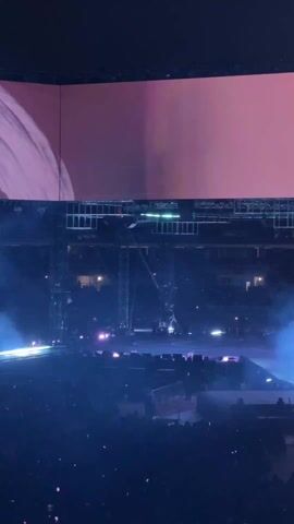 The video shows an entertainment event, possibly a concert, inside a large stadium. The scene is dominated by a large screen displaying a silhouetted figure. Below the screen, a stage is set up with spotlights and a red carpet. A large crowd is present, filling the seats of the stadium, and many people are holding up their phones. The lighting is predominantly blue, creating a dramatic atmosphere, and smoke or haze is present.