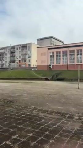 The video shows an outdoor, paved area, likely a courtyard or sports field, with a building in the background that has a light pink facade and multiple windows. There is a grassy slope behind a low retaining wall. There are some sparse trees and other buildings visible in the distance. A small, pink object appears to be dropped onto the paved area in the foreground. The overall impression is of a somewhat dreary, possibly neglected outdoor space.