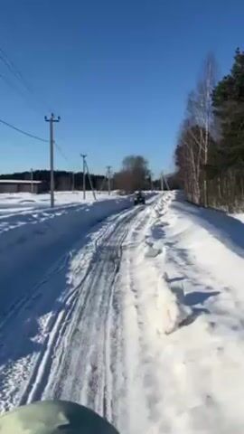 Here is a concise description of the video:

The video shows an ATV driving down a snowy path on a bright, sunny day. Power lines run parallel to the road, which is flanked by trees and snowdrifts. A building is visible in the distance.  The camera appears to be following the ATV.