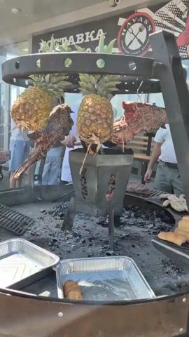 The video shows a close-up view of two pineapples and large cuts of meat roasting over an open fire. The pineapples are hanging upside down from a metal rack above the fire, with their leafy tops pointing downwards. The meat, which appears to be beef, is also hanging from the rack and is being exposed to the heat of the fire. There are people in the background, some of whom are looking at the roasting meat. The environment appears to be an outdoor cooking area with a grill and various cooking utensils visible. The scene is lively and suggests a communal gathering or event centered around barbecue.