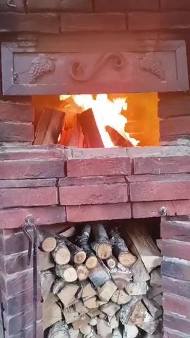 A close-up, stable shot shows a brick fireplace with a fire burning in the opening. There are logs of wood stacked neatly below the fire pit. The fire is bright and appears to be actively burning with flames consuming the logs. The decorative metalwork above the fire pit features grape vine motifs on the left and right, and a decorative scroll in the center. The overall scene suggests a cozy, outdoor setting.