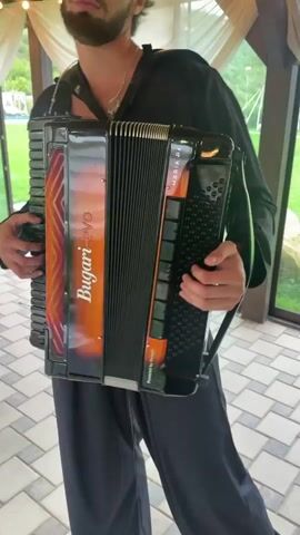 A man with a beard and dark curly hair, wearing a black shirt and pants, plays an accordion. He is standing outdoors under a tent, with a tiled patio and green grass visible in the background. He holds the accordion with both hands, his right hand on the keys and his left hand on the bellows, which he operates by expanding and contracting them. The accordion is black with orange accents and the brand name "Bugari evo" is visible on the side. The man occasionally looks towards the camera while playing.