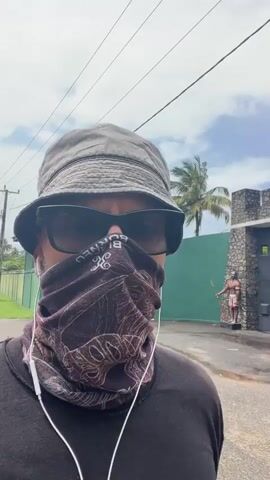 A first-person perspective video shows a person wearing a bucket hat, sunglasses, and a face mask, with earphones. In the background, there is a building with a large metal gate. To the right of the gate, there is a statue of a person holding a spear. The sky is partly cloudy.