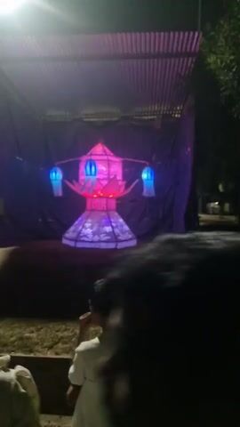 The video depicts a night scene in what appears to be a public park. There is a large, illuminated structure covered with a corrugated metal roof and blue and purple cloth in the background. This structure has decorative lighting displayed inside. The camera focuses on the structure and the surrounding environment, including patches of grass and scattered debris on the ground. Two people are visible in the foreground initially, and then two other men walk past the camera, briefly interacting with each other.  The lighting from street lamps and the illuminated structure provides the primary source of light in the dark environment.