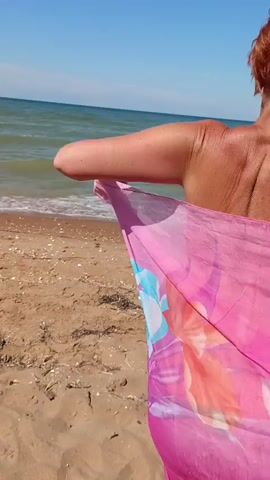 This media file captures a woman from behind, standing on a sandy beach with the ocean in the background. She has short, reddish-brown hair and is wearing a brightly colored sarong or wrap with a floral pattern in shades of pink, orange, and blue. The fabric is billowing in the wind, adding a sense of movement and freedom to the scene.

The setting appears to be a sunny day, with a clear blue sky and calm, green-blue water. The beach is sandy and appears to be relatively unpopulated, with some scattered debris like shells and dried seaweed along the shore. The sun is high in the sky, suggesting it is likely midday. The woman's posture, with her arms extended and the fabric flowing around her, conveys a feeling of joy and carefree energy. The overall impression is one of a relaxed and pleasant day at the beach.