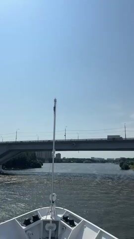 The video shows the bow of a white boat moving forward on a river. A tall mast with a black object at the top stands in the center of the bow. In the distance, a large concrete bridge spans the river. The sun is shining brightly in a clear blue sky. Trees and buildings are visible on the riverbanks. The water is rippling.