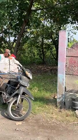A person wearing a hat and a colorful shirt is standing next to a cart attached to a motorcycle. The cart is filled with large white bags, some of which are tied with red and yellow strings. The motorcycle is parked on a dirt path in a grassy, wooded area. The person appears to be organizing or securing the bags in the cart. Sunlight filters through the trees, creating shadows on the ground.