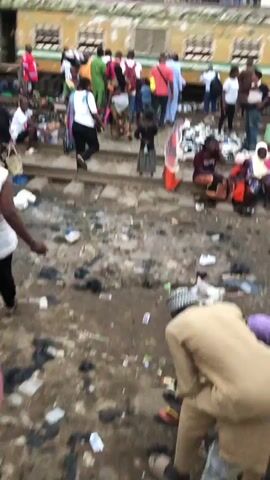 A group of people are gathered near train tracks. Many are dressed in casual clothing, while some wear traditional attire. There is a significant amount of trash and debris scattered on the ground, including plastic bags and other refuse. A train is partially visible in the background, and people are standing and sitting on or near the tracks. Some individuals appear to be waiting, while others are engaged in conversation or carrying bags. The overall scene suggests a busy, informal gathering or marketplace in an urban environment, with visible signs of litter and disarray.