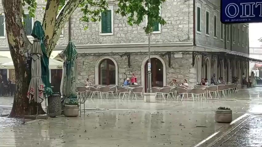 The video shows a rainy day in an outdoor cafe. The ground and tables are wet from the rain, and some of the umbrellas are closed. Several people are seated at the tables, some appearing to be eating or drinking. A large tree with a textured trunk is to the left of the frame, with closed umbrellas leaning against it. The cafe is attached to a stone building with arched windows and a green awning. A street lamp is visible in front of the building. A sign with Cyrillic lettering is visible above the cafe. The overall atmosphere is one of a quiet, somewhat deserted cafe during a downpour.