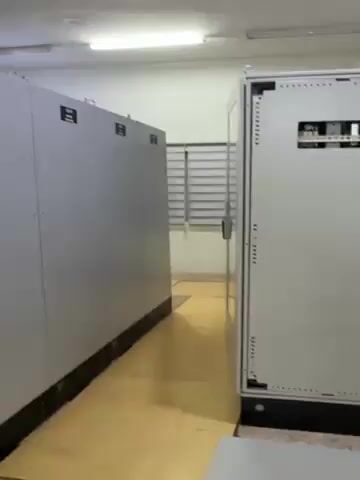 The video shows the inside of a control room with large gray electrical cabinets on one side. The door of one cabinet is opened, revealing a room with a large control panel along the back wall. A desk with a computer, books, and other equipment sits in the foreground. The room is lit by fluorescent lights. There are no people visible in the video.
