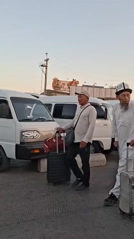 The video shows a street scene at dusk with several white minivans parked. A group of people is walking, some with luggage. An elderly man with a cane and a suitcase is in the foreground. A woman in a hijab pushes a stroller with a child. Another woman in a light-colored dress walks beside her. The sky is a pale orange and yellow.