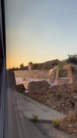 The video is taken from inside a moving vehicle, likely a bus, looking out the window. The time of day appears to be sunset, with the sky displaying warm orange and yellow hues. Outside the window, a roadside scene unfolds. A road runs horizontally across the frame, with vehicles, including what appear to be vans and possibly a truck, traveling on it. The landscape consists of a rocky, hilly terrain with sparse vegetation. There are some concrete structures or barriers visible along the road and on the embankment. The sun is low on the horizon, casting a warm glow on the scene. The interior of the vehicle is visible on the left side of the frame, showing seats and window coverings. The perspective suggests a journey along a highway or rural road.