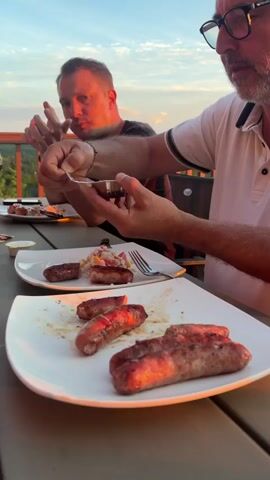 The video shows two men having dinner outdoors on a deck. In the foreground, a plate of grilled sausages is visible. A man with glasses is holding a fork and knife, appearing to cut into a sausage. Another man is seated across from him, looking at the camera and gesturing with his left hand. The background reveals a scenic view of hills and trees bathed in the warm light of a sunset, with a clear blue sky above. The scene conveys a relaxed outdoor dining experience.