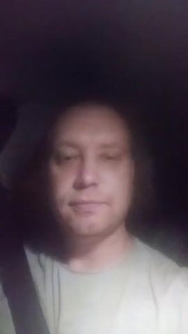 The video begins with a close-up, head-and-shoulders shot of a man with medium-length, dark, wavy hair. He is wearing a light-colored t-shirt and a seatbelt is visible across his chest. The background is dark, suggesting the shot was taken at night or in a dimly lit environment, likely inside a vehicle given the seatbelt. The man's face is the primary focus, and his expression is neutral. As the video progresses, his mouth opens as if speaking, though no audio is provided in the description. The lighting is soft, casting a slight shadow on his face, and his eyes appear to be looking directly at the camera. The overall impression is of a person in a car, possibly recording a message or talking to someone.