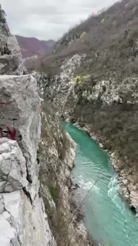 The video shows a wide turquoise river flowing through a rocky canyon. The river is surrounded by steep, rocky cliffs covered with sparse vegetation. The sky is overcast and the lighting is somewhat muted. The camera pans slowly to follow the course of the river, revealing its winding path through the canyon. There are no people visible in the frame.