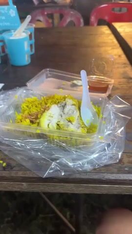 The video shows a close-up of a takeout container filled with yellow rice and chicken, placed on a wooden table. A white plastic spoon is resting in the container. To the right of the container, there is a small plastic cup with a red sauce. In the background, out of focus, are several blue cups with white spoons, and beyond that, a bustling night market scene with people, stalls, and lights. A large fan is visible in the middle ground. The lighting suggests it is nighttime.