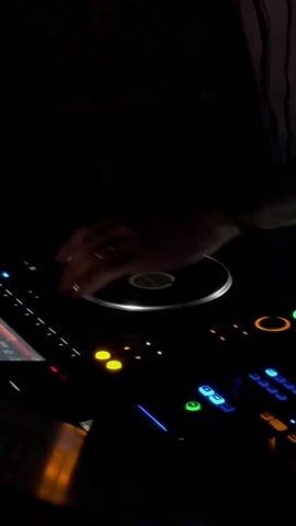 Here's a description of the video:

A DJ is shown from a close, low angle, operating a DJ controller in a dimly lit environment, presumably a nightclub. The DJ, wearing a striped shirt, manipulates the turntable and touches various buttons and controls on the device. The DJ controller is illuminated with a variety of bright, colorful lights, including blue, yellow, green, and red, highlighting its features. A small screen on the controller is visible, displaying some kind of audio information. The background is mostly dark.
