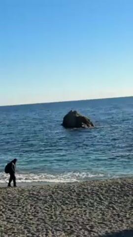 The video opens on a beach with dark sand and rolling waves on a clear, sunny day. In the distance, a rocky outcrop is visible in the sea. The camera then pans to the left to show a statue on top of a large rock formation. The statue appears to be a figure kneeling with its head bowed, supporting a large beam or cross. The sun is shining behind the statue, creating a silhouette effect. A bird flies across the frame. The sea is visible in the background.
