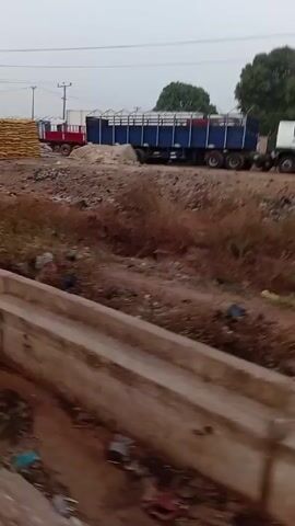 The video, taken from a moving vehicle, presents a scene featuring industrial activity near a roadside.

Initially, large trucks are visible on a dusty, uneven ground alongside the road. One truck is blue and appears loaded with cargo, while another, smaller red truck is positioned nearby. There are also large piles of yellow goods, possibly packaged or wrapped. In the background, power lines stretch across an overcast sky, and some green trees are present.

As the video progresses, the perspective shifts downwards, focusing on a concrete drainage ditch running parallel to the road. The ditch is filled with dirty water and accumulated trash, highlighting the environment's neglected state.