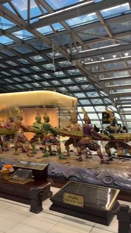 The video shows a large, ornate sculpture at an airport. The sculpture depicts figures pulling on what appears to be a giant serpent or rope. In the foreground, there are several individuals, some of whom are wearing traditional clothing and headdresses. They are shown in a pose that suggests they are engaged in a collective effort. The main sculpture is topped by a multi-armed purple deity standing on a rocky formation. Surrounding the base of the sculpture are representations of water and waves, as well as a large turtle. The environment is clearly indoors, with a high, glass-paneled ceiling and visible airport signage and infrastructure in the background, including escalators and carts. There are also smaller decorative elements like flower arrangements. The lighting is bright, suggesting artificial illumination or natural light filtering through the ceiling.