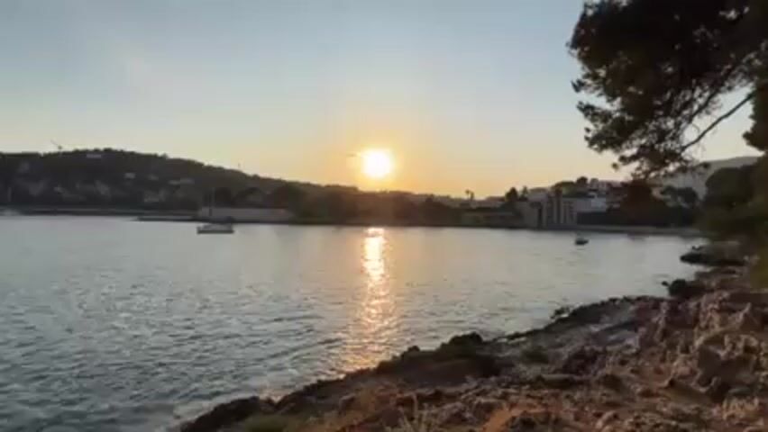 Here is a detailed description of the video:

The video begins with a scenic shot of a sunlit ocean at sunset. In the distance, buildings and trees line the coast, while a few boats dot the water. The foreground features rocky terrain with some vegetation. The sun's reflection creates a bright streak on the water's surface. 

The camera then rotates to show a couple lying down together, facing the camera. The woman wears sunglasses and a blue top, while the man also has sunglasses and a light-colored shirt. They both have their heads close together and are looking directly at the camera.