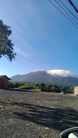 The video shows a mountain with clouds partially obscuring its peak, viewed from a clearing with gravel on the ground. A small, brown wooden structure is visible to the left of the frame, along with a tall transmission tower behind it. A leafy tree stands in the foreground to the left. Overhead power lines cross the sky. The overall scene is outdoors under a clear blue sky.